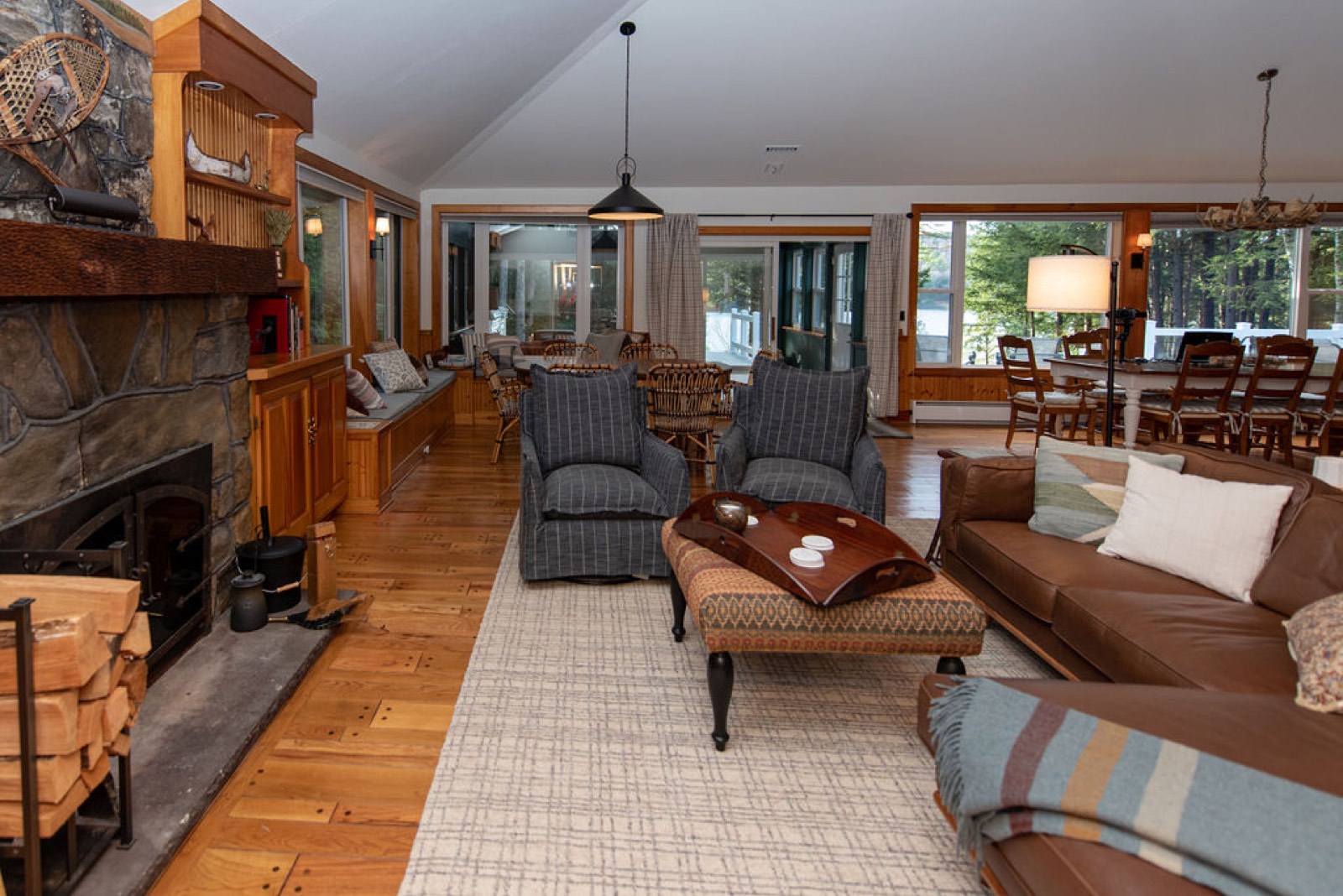The great room at Camp Sunnyside with a fieldstone fireplace stacked with firewood, honey-pine floors, leather seating, and a wall of glass to the pines.